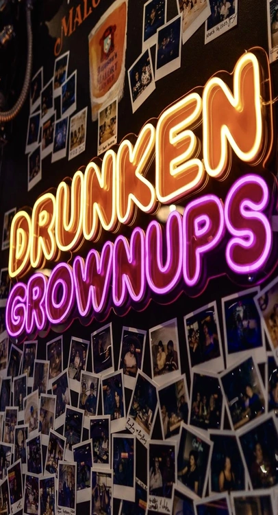 Neon sign reading “Drunken Grownups” glowing in yellow and purple, mounted over a wall covered in polaroid photos.