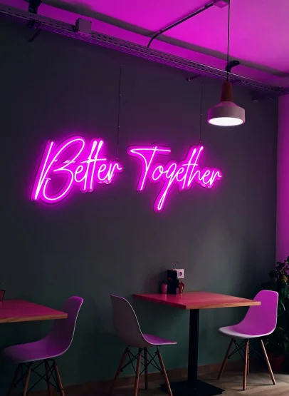 Pink neon sign reading “Better Together” mounted on a café wall above a small table and chairs.